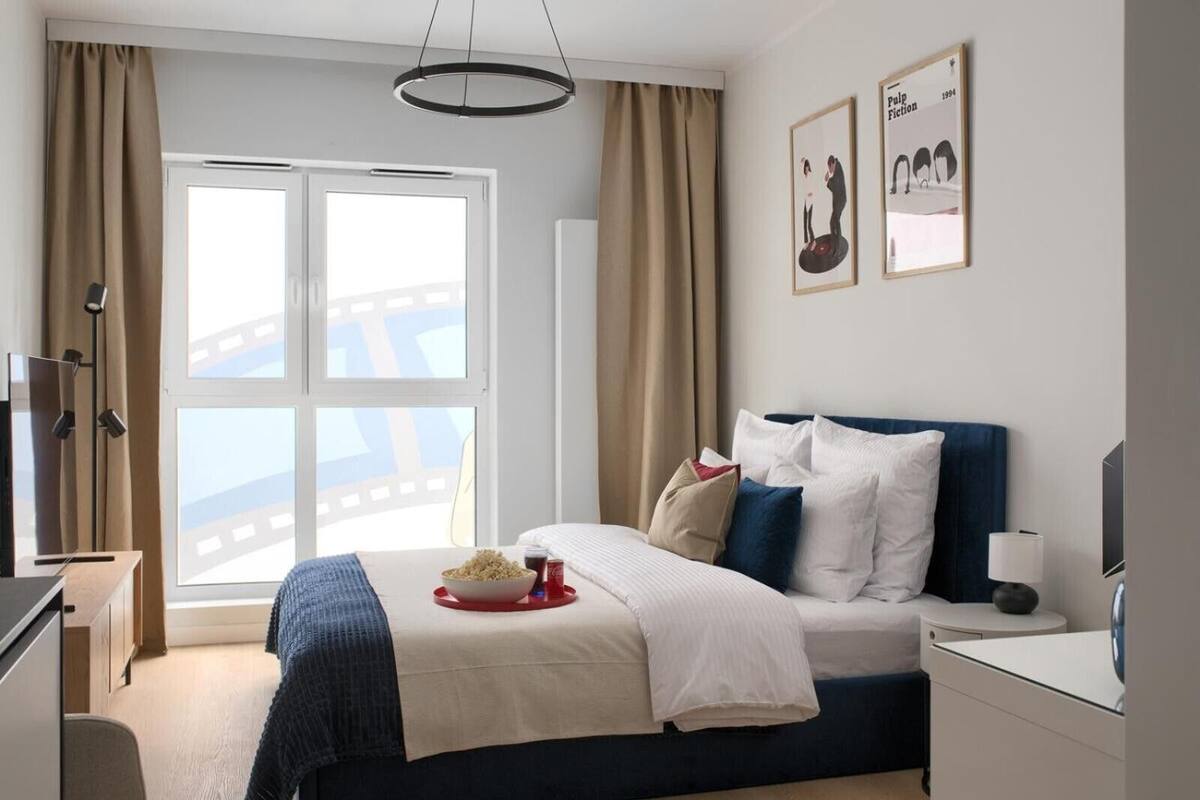 Une chambre d’hôtel comprenant un lit, un bureau, une chaise, une fenêtre avec des rideaux et un mur orné de tableaux.