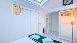 Une chambre avec un puits de lumière, un lit recouvert d’une couverture verte, une armoire et un placard où sont suspendus des vêtements.