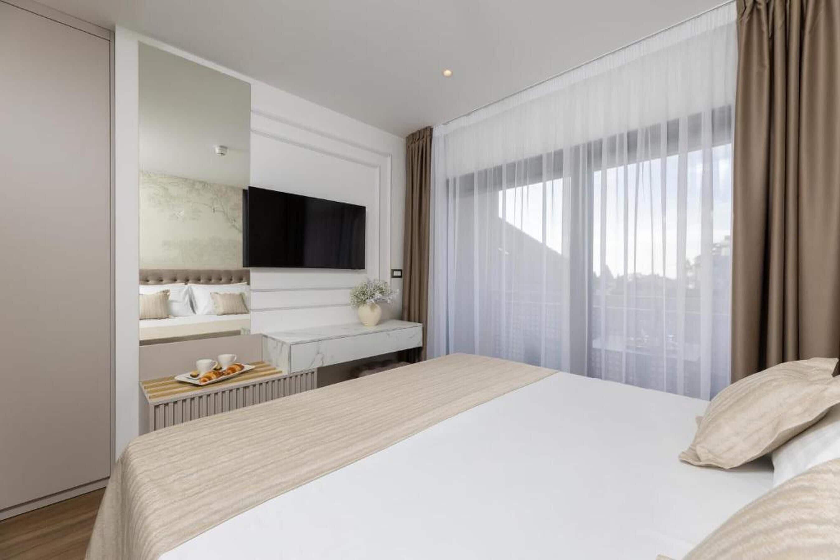 A modern hotel room with a large bed, a flat-screen TV mounted on the wall, a small table with a plant, and a window with sheer curtains.