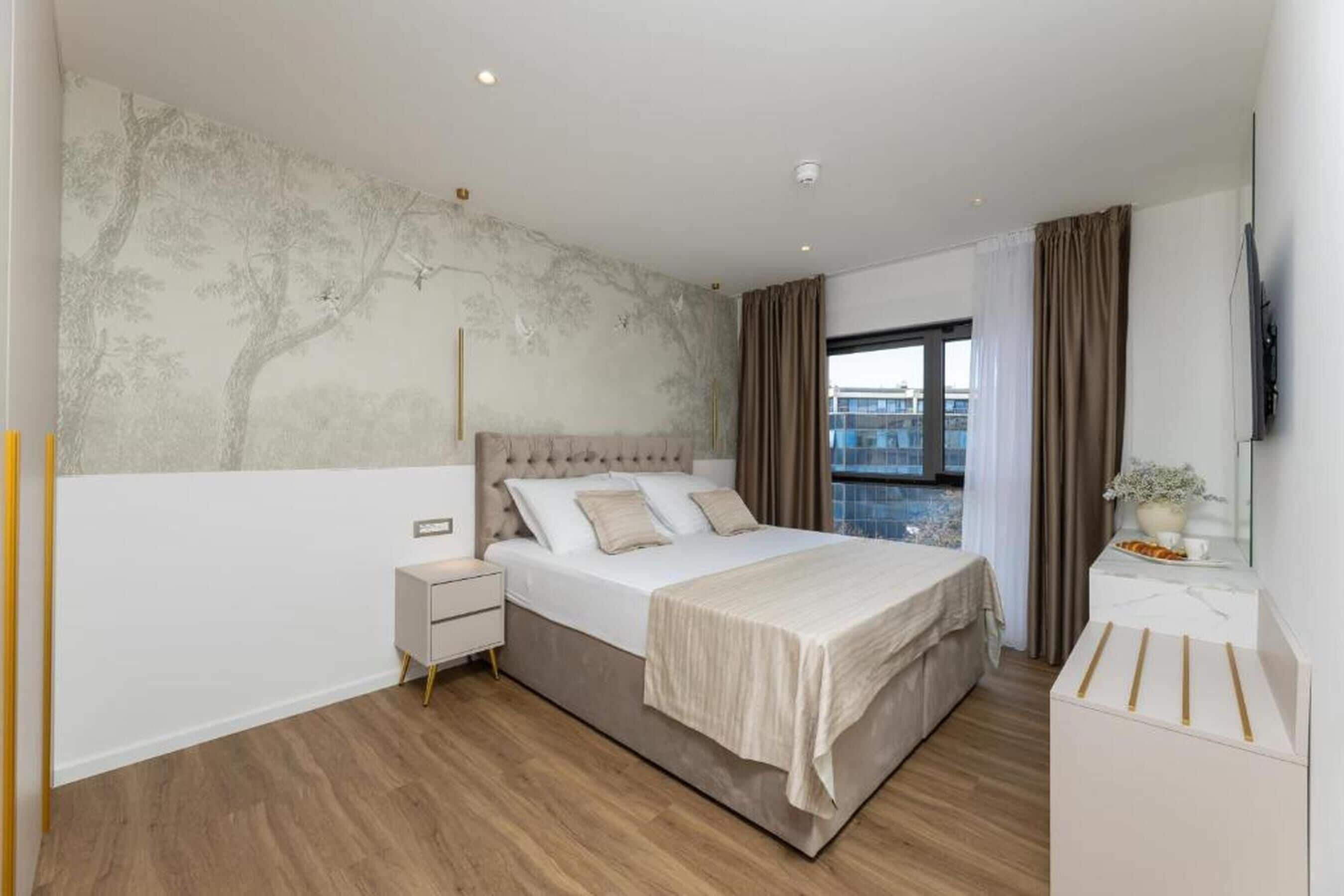 A modern bedroom with a large bed, a nightstand, a TV, and a view of the city.