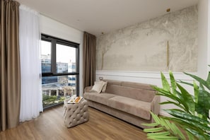 A modern living room with a beige sofa, a tufted ottoman, and a large window with curtains. - Sunday Residence and SPA Split (Split)