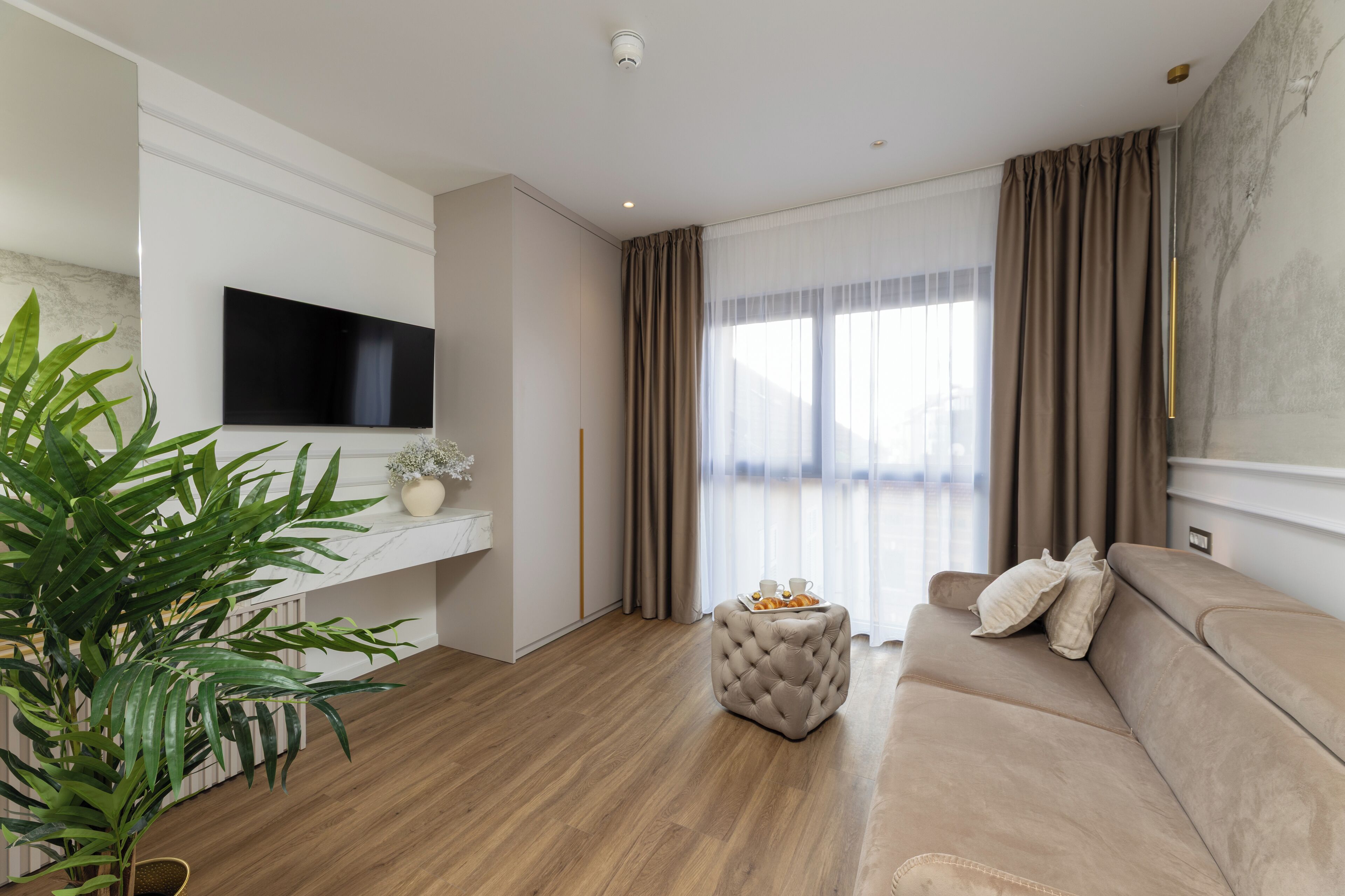 A modern living room with a flat-screen TV, a beige sofa, a tufted ottoman, and a large plant.