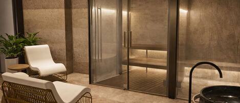 A modern spa area with a glass-enclosed sauna, two lounge chairs, a round wooden bench, and a large black basin.