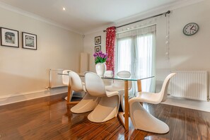 A modern dining area with a glass table and white chairs, a vase with purple flowers, and a clock on the wall. - Links Lodge | Blacklaws Steading (Anstruthe)