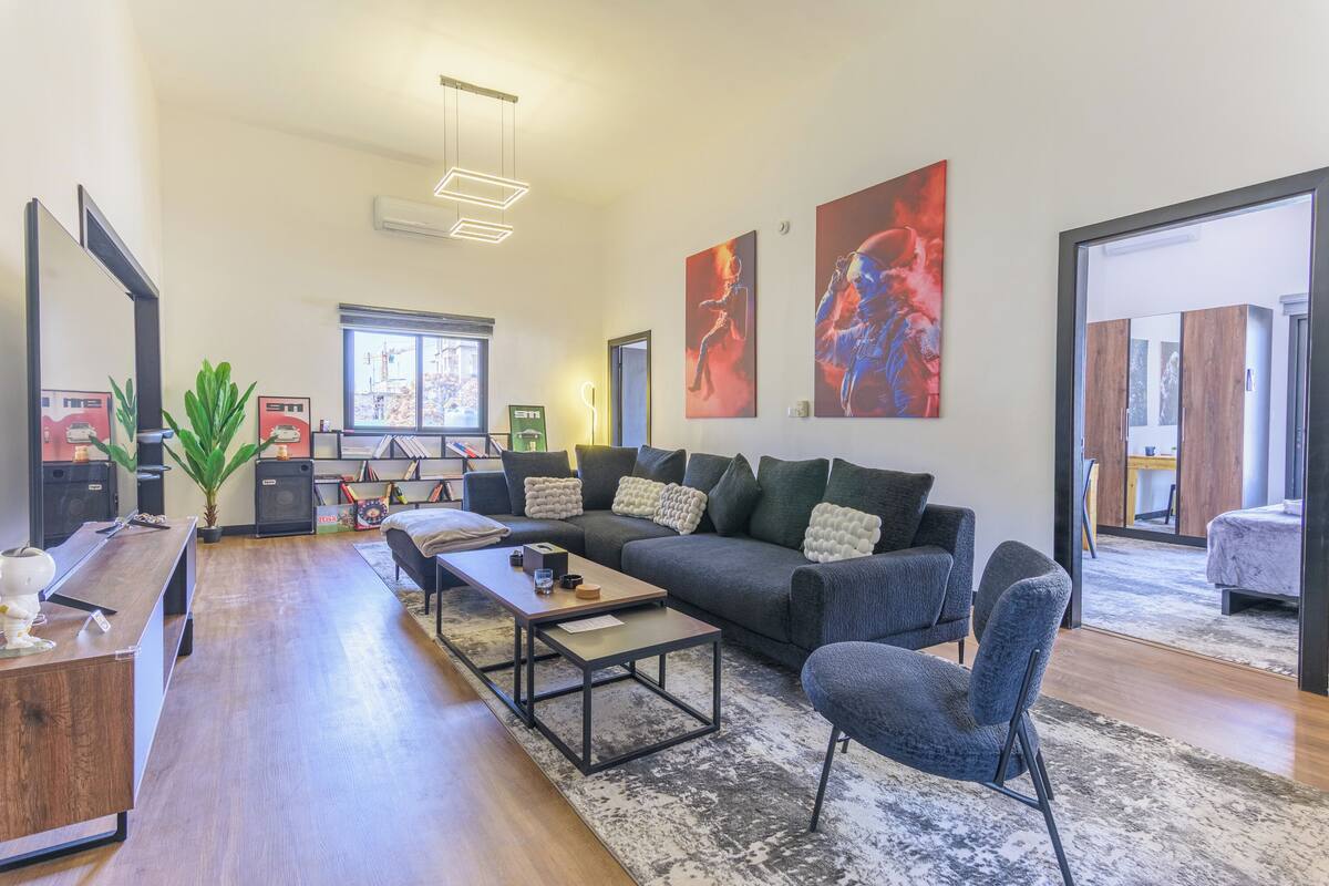A modern living room with a dark grey sofa, a wooden coffee table, and a grey armchair. There are abstract paintings on the walls, a large window, and a door leading to another room.