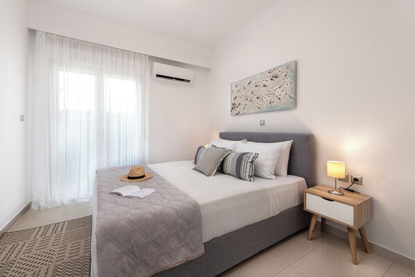 A bedroom with a bed, bedside table, lamp, and a wall-mounted air conditioner.
