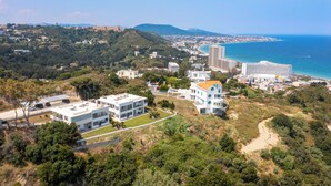 Vue aérienne d’une zone côtière avec des bâtiments modernes, une plage et un paysage montagneux en arrière-plan.