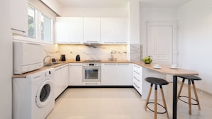 Una cocina moderna con armarios blancos, horno empotrado, lavadora y una mesa de comedor de madera con dos taburetes.