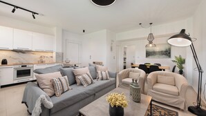 Un salon moderne avec un canapé gris, des fauteuils beige, une table basse ornée d’un vase, et un coin repas comprenant une table et des chaises.