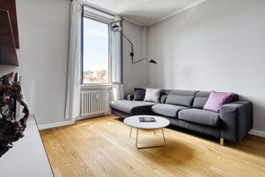 En moderne stue med en grå sofa, et rundt sofabord, en gulvlampe og et stort vindue med udsigt til bygninger.