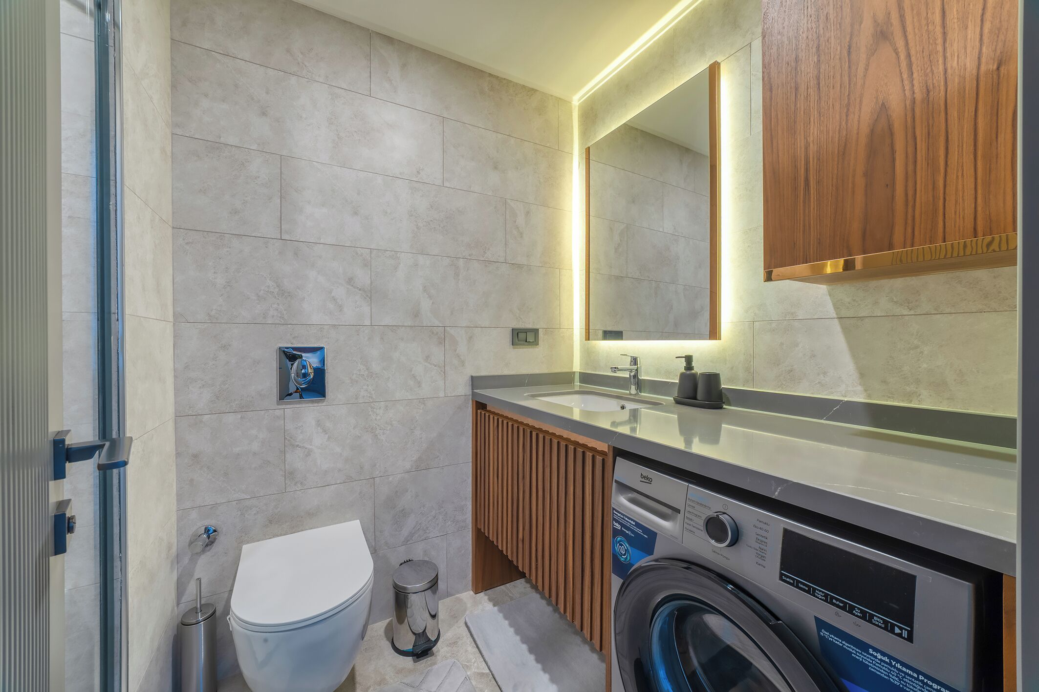 Une salle de bain moderne équipée de toilettes, d’un lave-linge et d’un miroir au-dessus du lavabo, avec un meuble en bois.