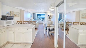 Une cuisine moderne et compacte, dotée de meubles blancs et d’un four à micro-ondes. On aperçoit un coin repas avec une table et des chaises, qui donne sur un balcon offrant une vue sur l’océan.