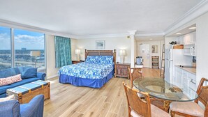 Habitación de hotel con cama, sofá, mesa de comedor con sillas, zona de cocina y un amplio ventanal.