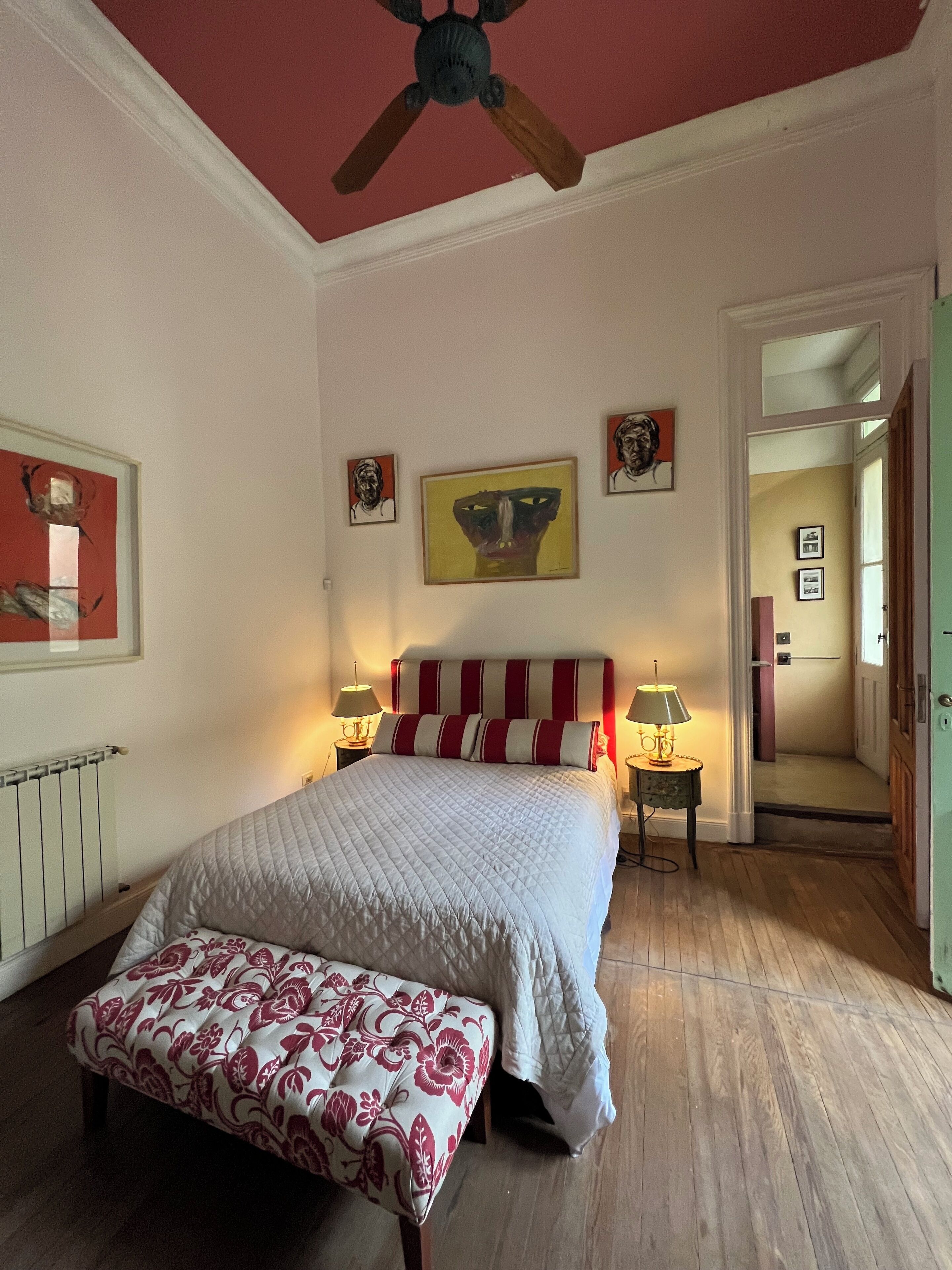 A bedroom with a bed, a bench, a ceiling fan, and artwork on the walls.