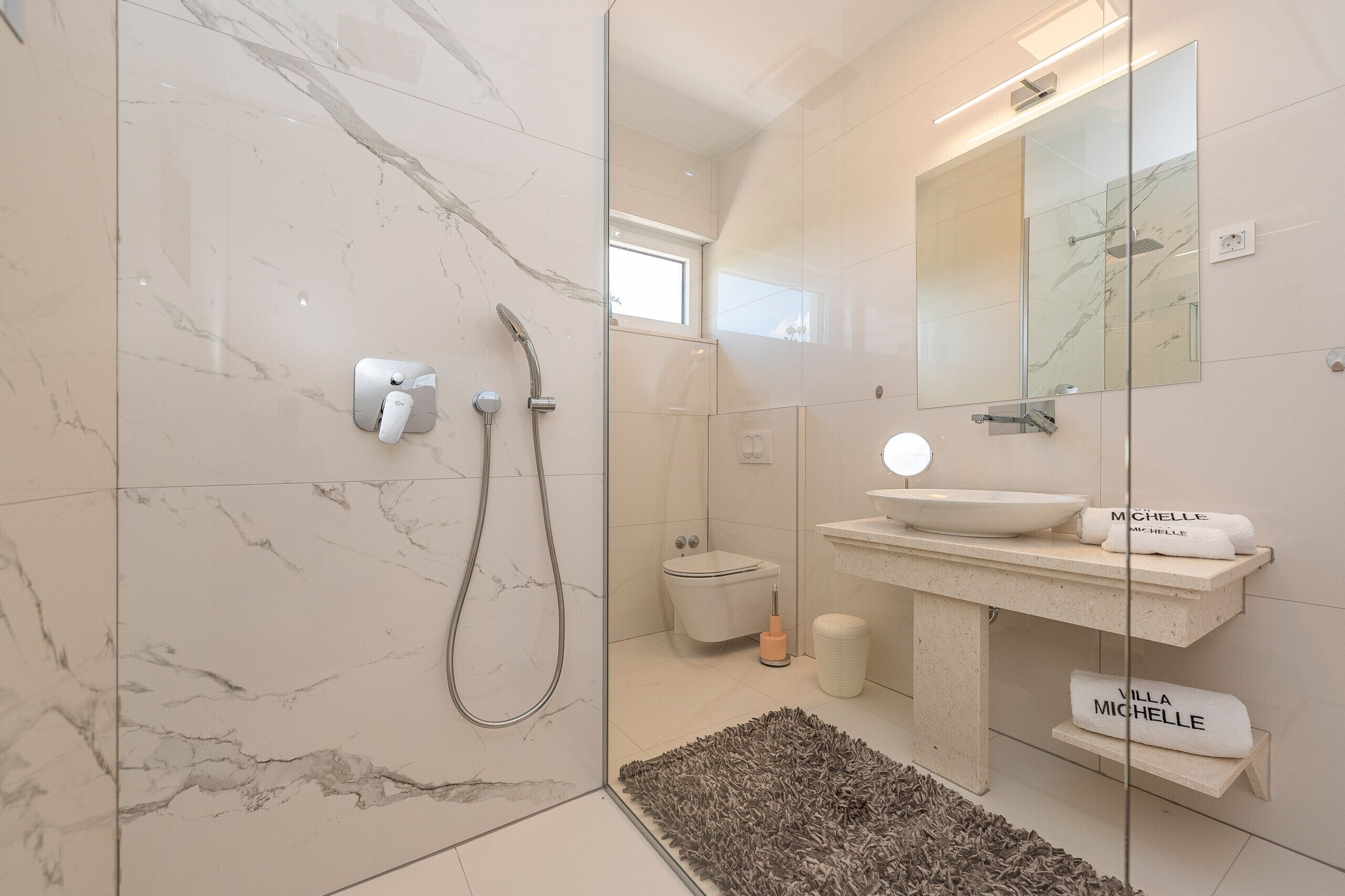 A modern bathroom with a glass-enclosed shower, a white marble floor, a sink with a mirror above, and towels labeled "Villa Michele".
