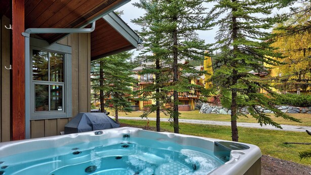 A hot tub outside a building with a view of a forest and a building in the background. - Spacious, Multi-Floor 4bedroom Townhome Walking Distance to Village, Along Orient Chairlift! (Sun Peaks)