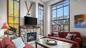 A cozy living room with a stone fireplace, large windows, a television, and a red sofa. - Spacious, Multi-Floor 4bedroom Townhome Walking Distance to Village, Along Orient Chairlift! (Sun Peaks)