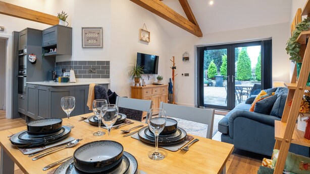 A modern kitchen with grey cabinets, a dining table set for six with black plates and wine glasses, and a blue sofa in the living area. - Beautiful 2 Bedroom Barn in Cerney Wick (Cirencester)