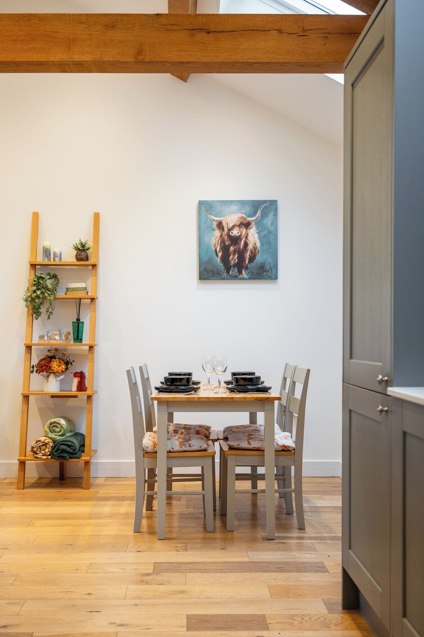 A dining area with a wooden table and chairs, a wooden ladder shelf with various items, and a painting of a yak on the wall.