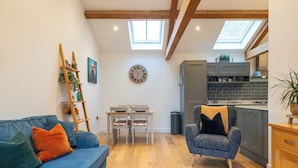 A cozy living room with a blue sofa, a small dining table with chairs, a grey armchair, and a kitchen area with grey cabinets and a black tiled backsplash. - Beautiful 2 Bedroom Barn in Cerney Wick (Cirencester)