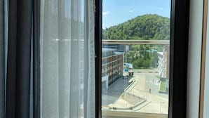 A view through a window with a curtain, revealing a building complex with a green hill in the background. - Lovely 1-bedroom apartment in Bakuriani perfect for a cozy getaway (Bakuriani)