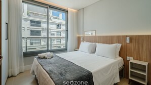 A hotel room with a large window, a bed, a bedside table, and a wooden headboard. -  Apt w/ Barbecue Grill 50 m from the Beach DOI0203 (Florianópolis)