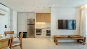 A modern kitchen with a built-in oven, microwave, and refrigerator. There is a dining area with a wooden table and chairs, and a TV mounted on the wall. -  Apt w/ Barbecue Grill 50 m from the Beach DOI0203 (Florianópolis)