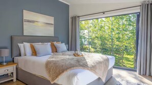 A modern bedroom with a large bed, a bedside table, a nightstand, and a view of greenery through the window. - Isca Lodge - With Decking (York)