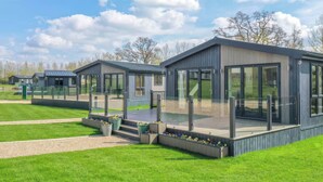 A modern, single-story building with large windows and a flat roof, surrounded by well-maintained grass and flower beds. - Isca Lodge - With Decking (York)