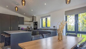 A modern kitchen with dark cabinets, a central island, and a dining area with a wooden table and leather chairs. - Isca Lodge - With Decking (York)