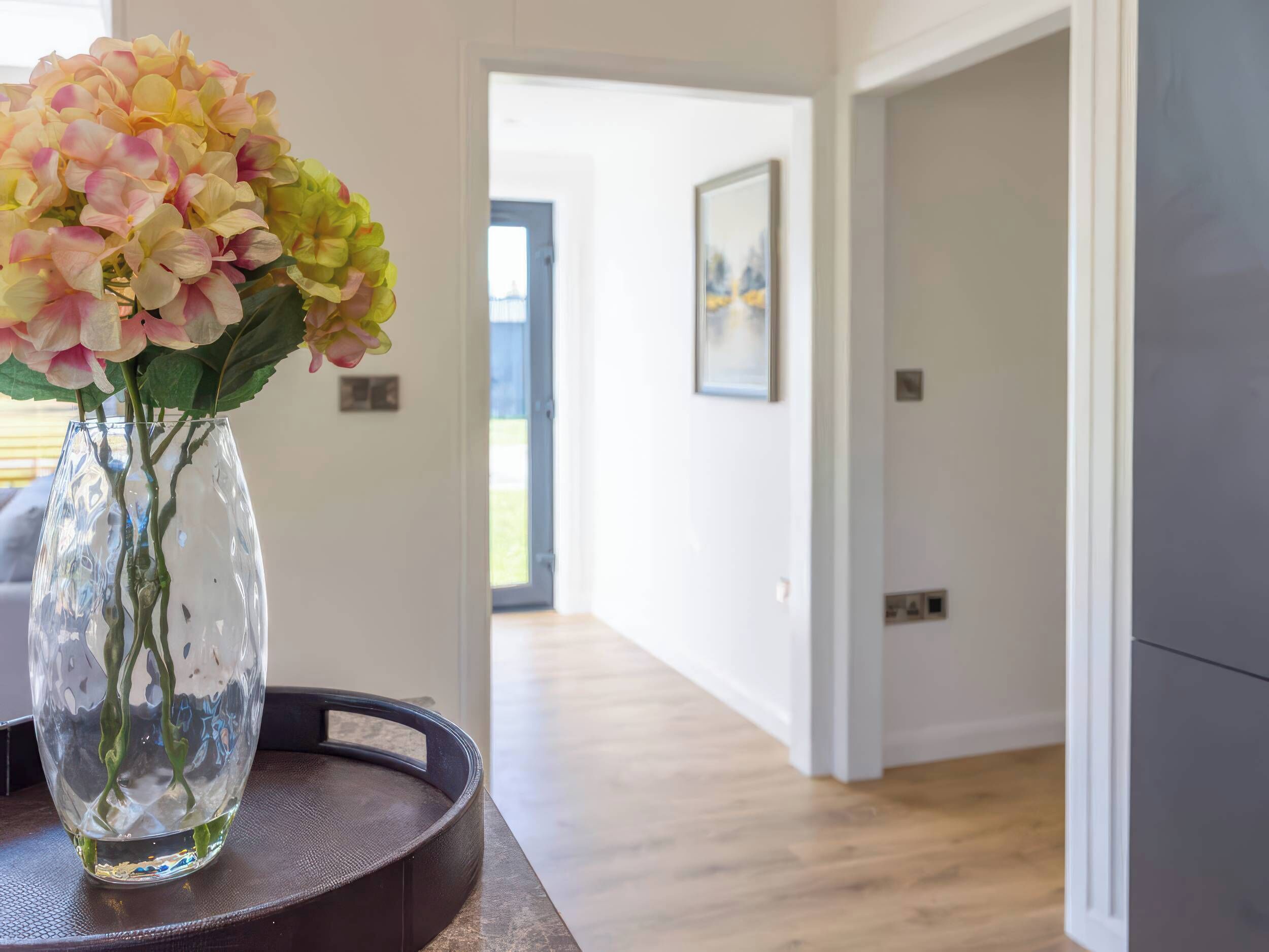 On voit clairement une pièce avec un vase de fleurs posé sur une table, une porte donnant sur une autre pièce et un tableau encadré accroché au mur.