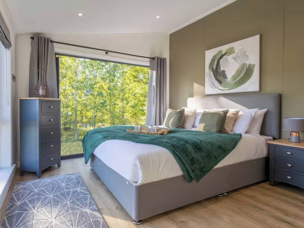 A modern bedroom with a large bed, a dresser, a nightstand, and a view of trees through a window. - Isca Lodge - With Decking (York)