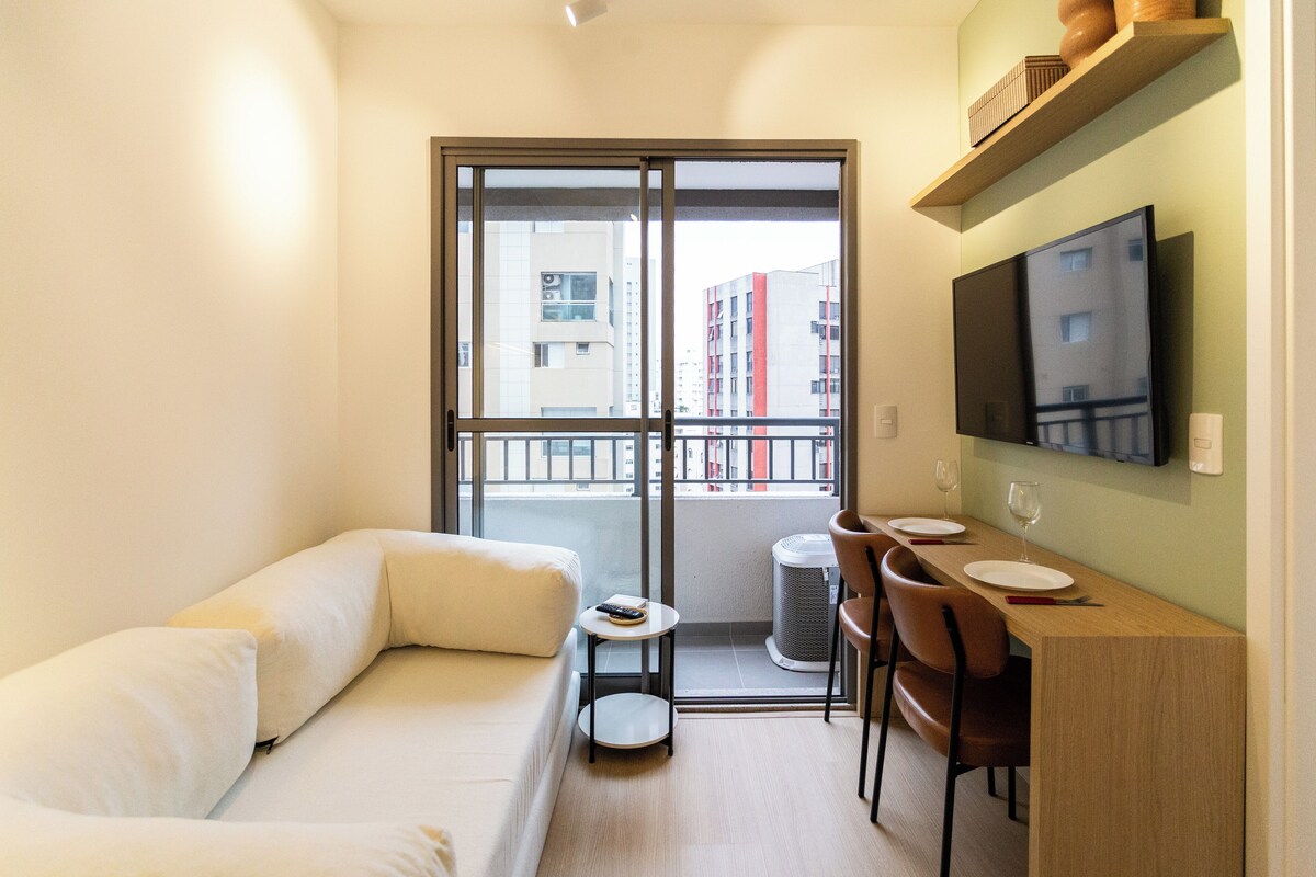 Un espacio habitable moderno y compacto con sofá, una mesita con un control remoto, una zona de comedor con sillas y una mesa, y un televisor montado en la pared.