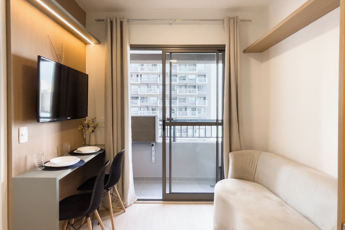 Una sala moderna con un televisor de pantalla plana, un comedor con mesa y sillas, y un ventanal corredero que da a un balcón con vistas a un edificio residencial.