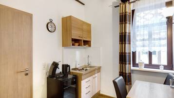 A compact kitchen with a sink, stove, and cabinets, a dining table with chairs, and a window with curtains.