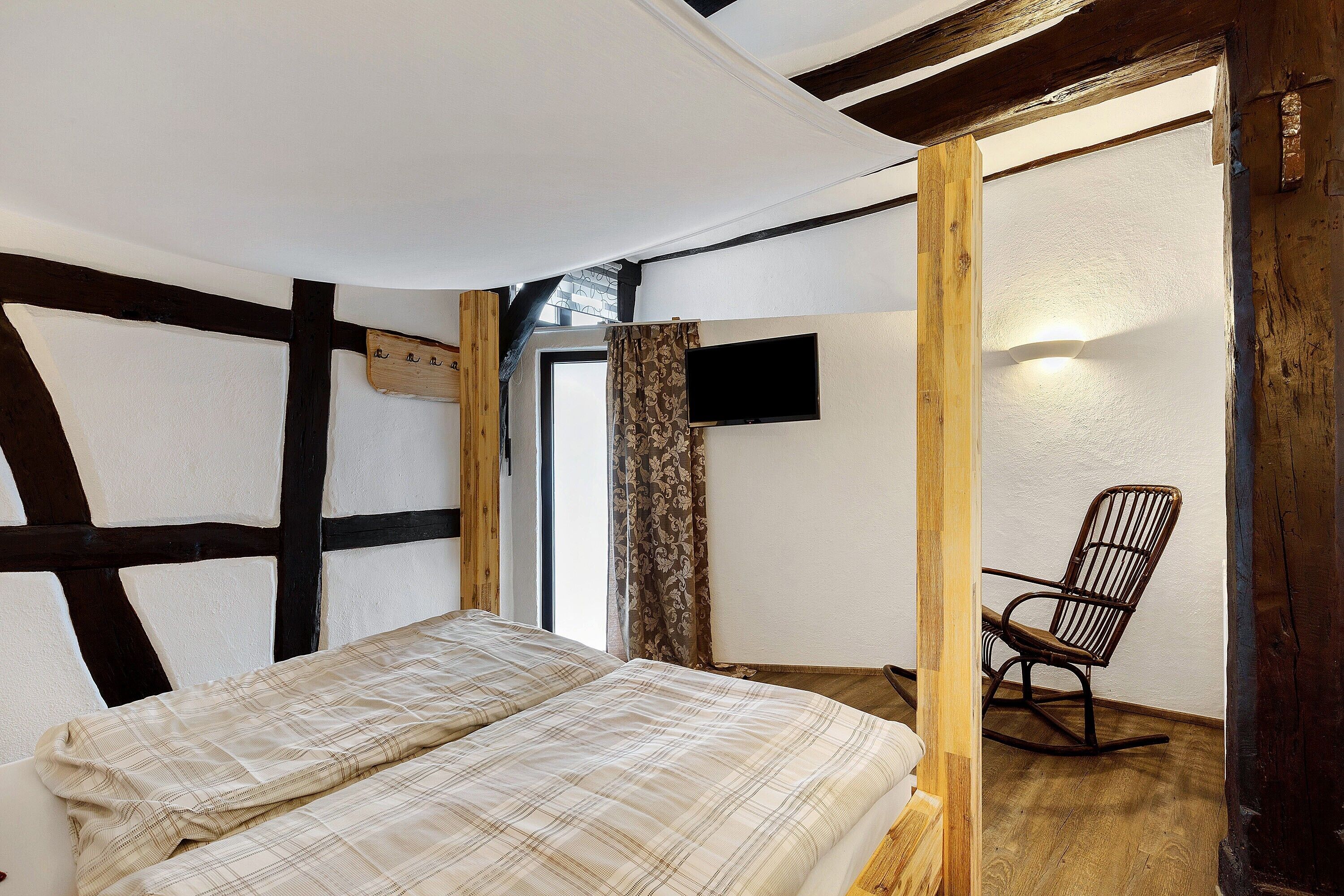 A room with exposed wooden beams, a bed with a plaid bedspread, a rocking chair, and a television mounted on the wall.