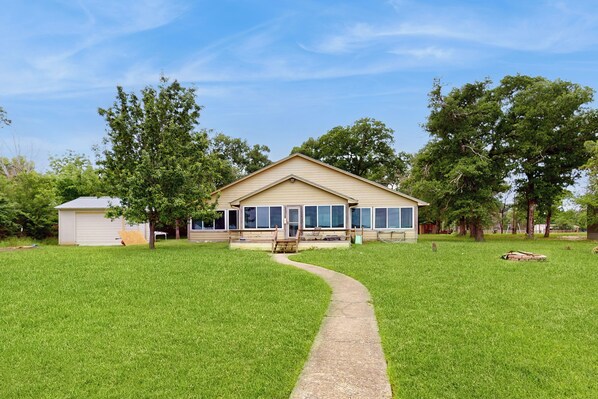 A single-story house with a front porch, a small shed to the left, and a paved driveway leading to the entrance.