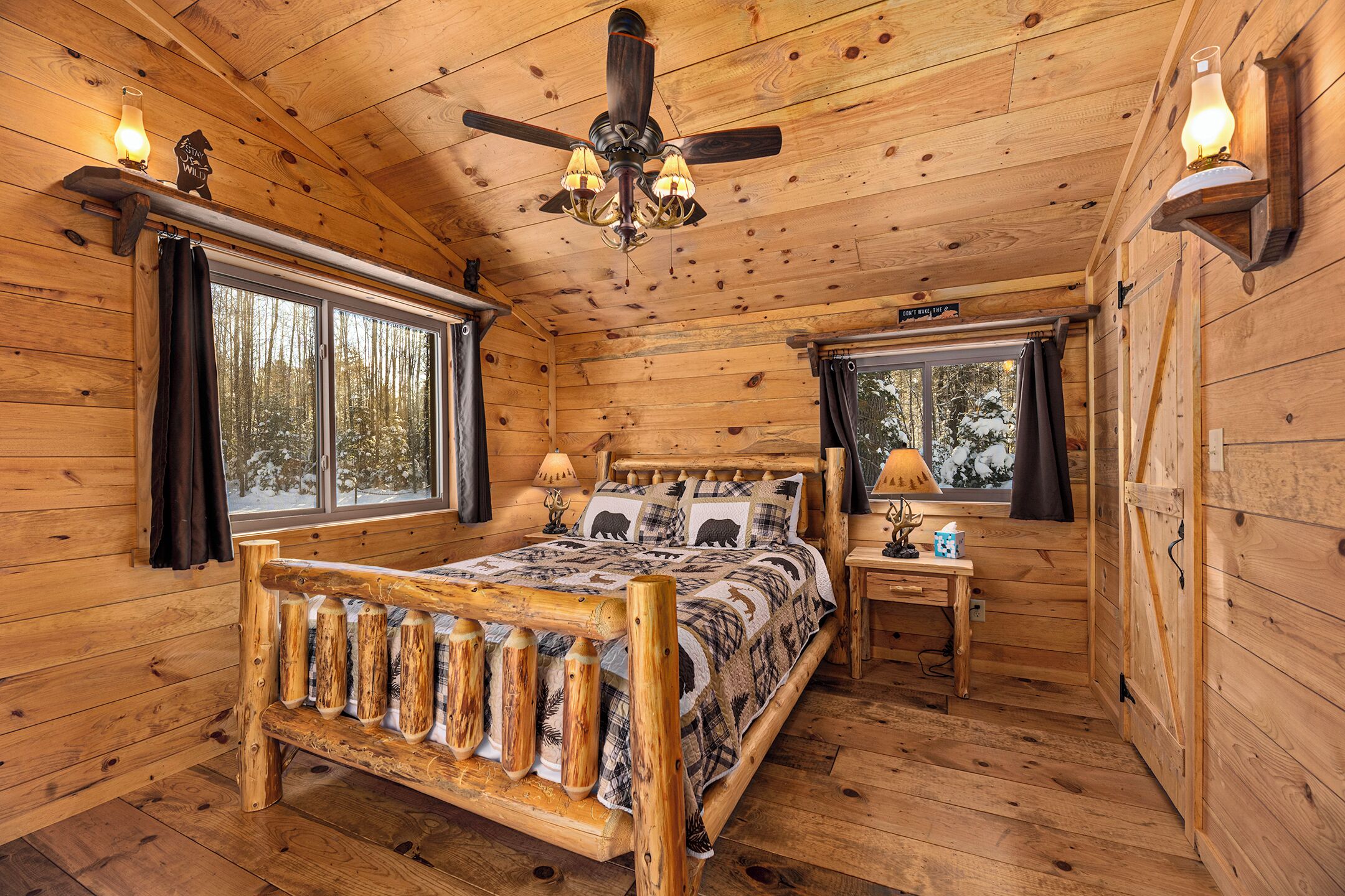 Ein Schlafzimmer einer Holzhütte mit einem Bett, zwei Fenstern, einem Deckenventilator und einem Nachttisch mit Lampe.