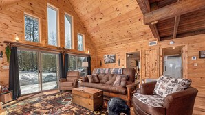 A log cabin interior with a large window, a sofa, a chair, a wooden chest, and a black dog. - Hot Tub, Firepit, AC, Near Trails & Lakes (Phelps)