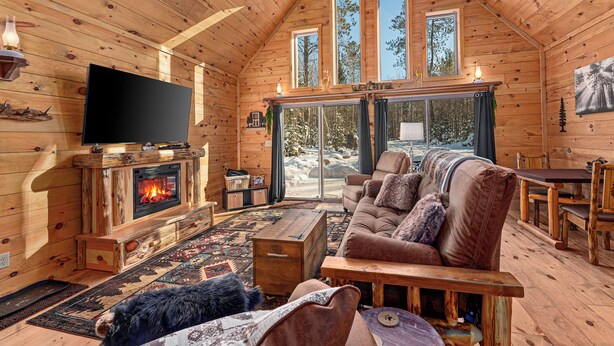 A cozy log cabin interior with a fireplace, a large TV, a comfortable sofa, and a dining area. - Hot Tub, Firepit, AC, Near Trails & Lakes (Phelps)