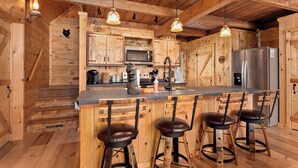 A wooden cabin kitchen with a central island, bar stools, a microwave, a refrigerator, and hanging pendant lights. - Hot Tub, Firepit, AC, Near Trails & Lakes (Phelps)