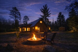 A rustic cabin with a fire pit, outdoor seating, and a view of the surrounding forest at dusk.