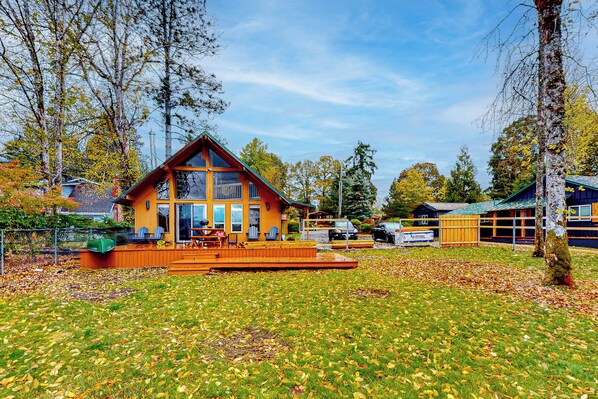 A two-story wooden cabin with a deck, surrounded by trees and a grassy yard. - 2 BR Lakeside Home w/ Wood-burning Fireplace (Silver Lake)