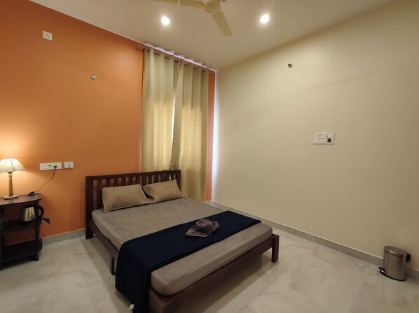 A bedroom with a bed, a nightstand, a lamp, a ceiling fan, and a window with curtains. - TADY Homes (Tiruvannamalai)