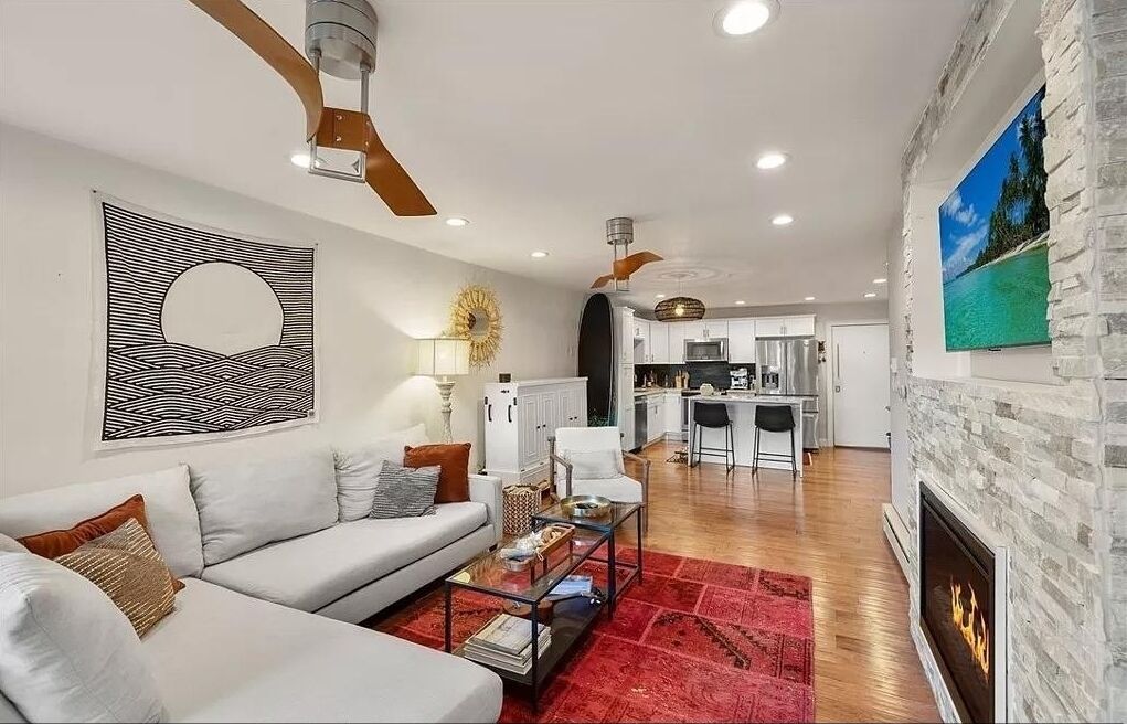 A living room with a sofa, coffee table, and fireplace. There is a kitchen in the background with modern appliances.