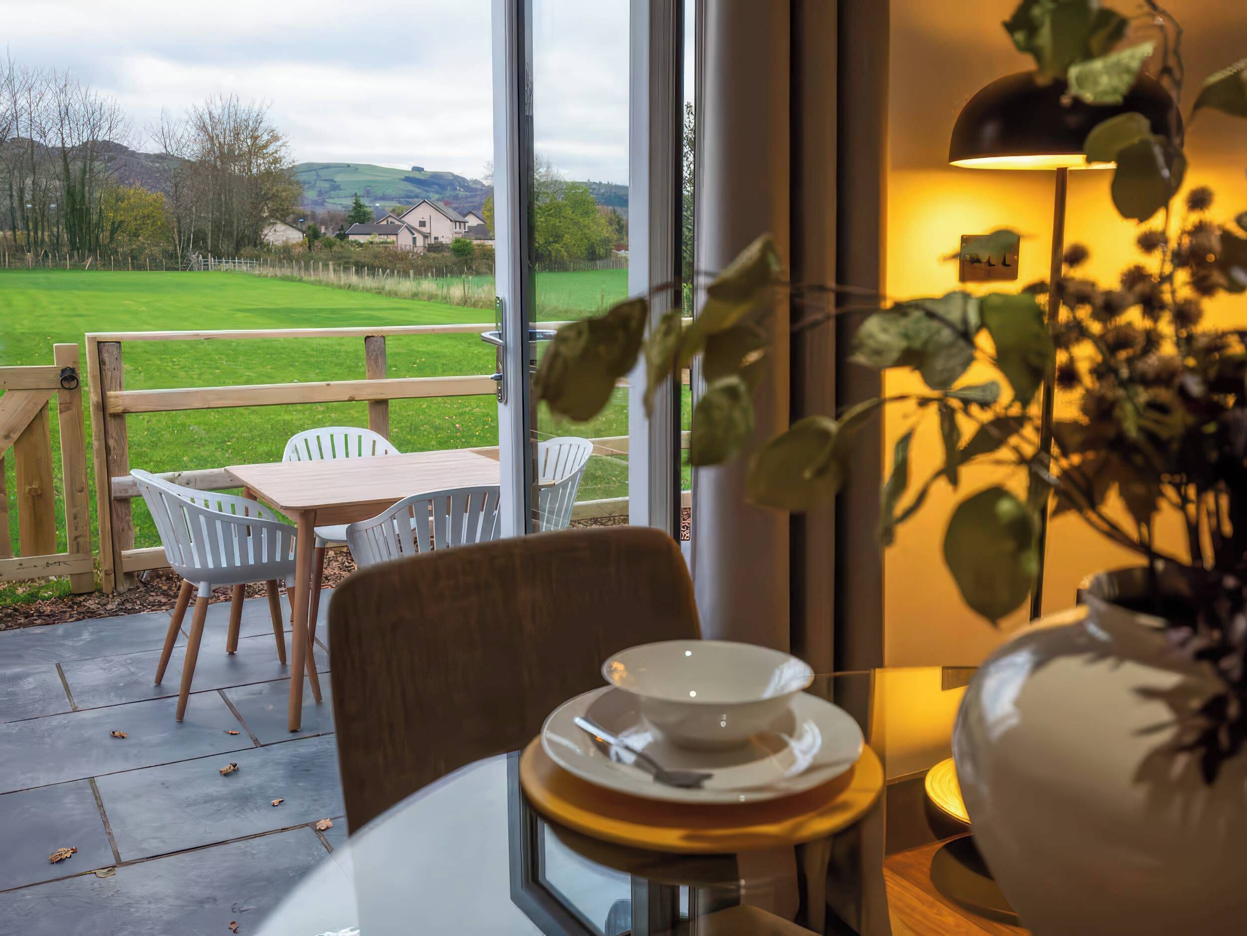 Un coin repas avec une table et des chaises, une vue sur un champ verdoyant et des maisons, et une plante en pot.