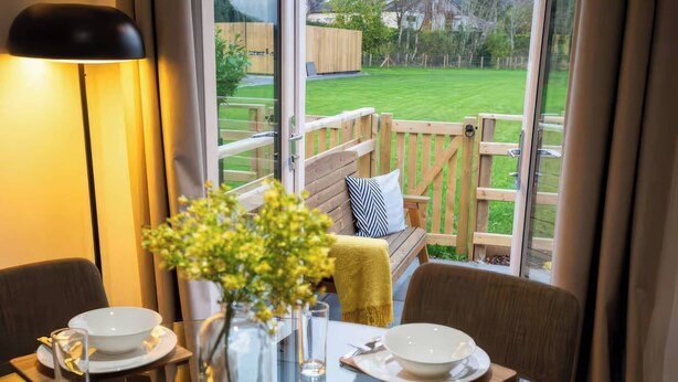 A dining table set with plates, glasses, and a vase with flowers, overlooking a wooden deck and a grassy yard. - Fir Cottages (Llanrwst)