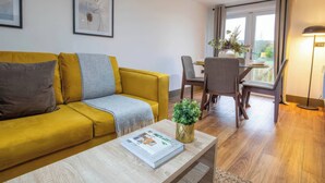 A modern living room with a yellow sofa, a coffee table with a magazine, and a dining area with a table and chairs. - Fir Cottages (Llanrwst)