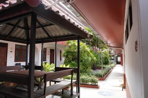 Un espacio de estar al aire libre cubierto con una mesa y bancos, rodeado de plantas en macetas y un sendero que lleva a un edificio.