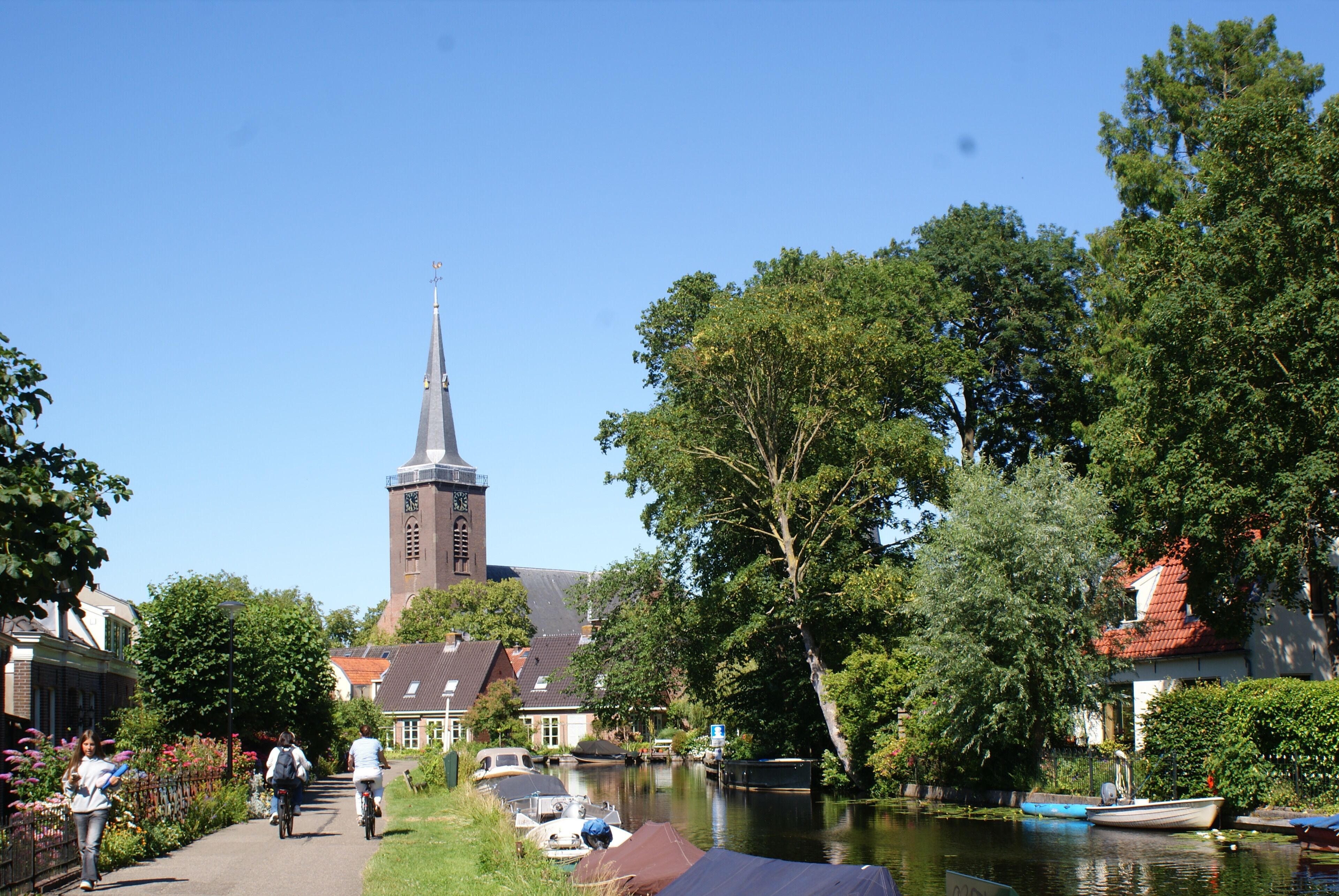Een gracht met boten, een kerk met een hoge toren en een voetpad.